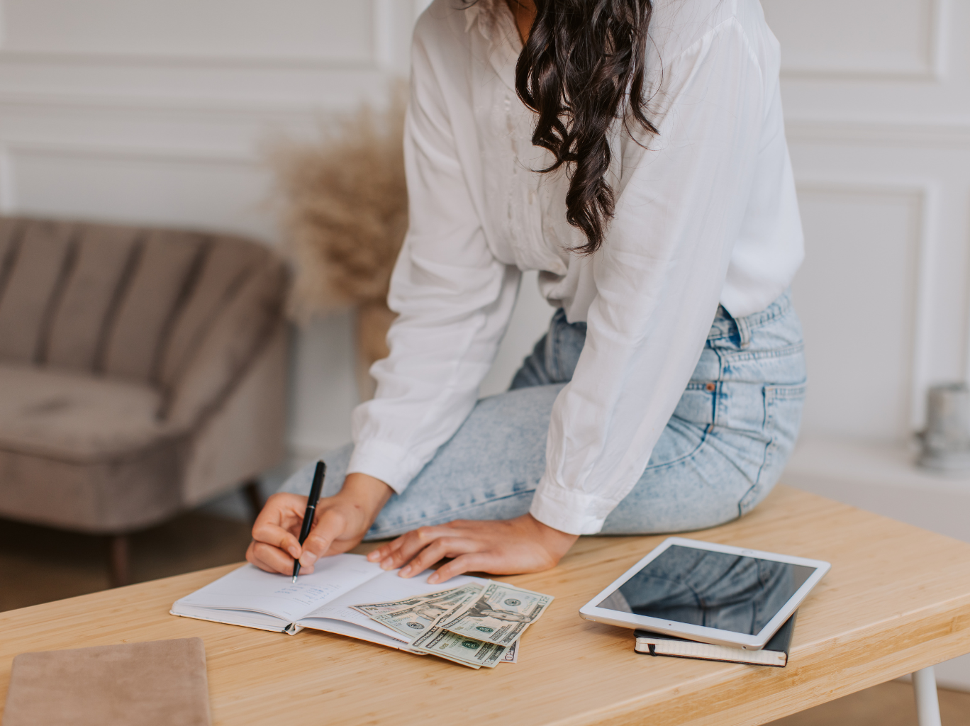 Female entrepreneur sitting on the ledge of a table, counting cash, writing in her notebook and learning how to leverage compound interest to maximize wealth and financial growth.