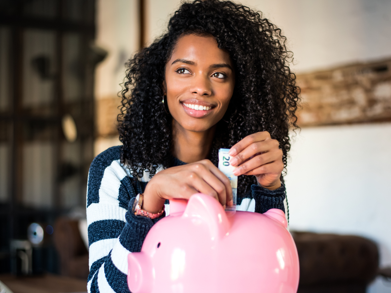 A young black female entrepreneur sitting in an office at the bank with a piggy bank on the desk. She holds money in her hands and is looking out and contemplating her business finances.
